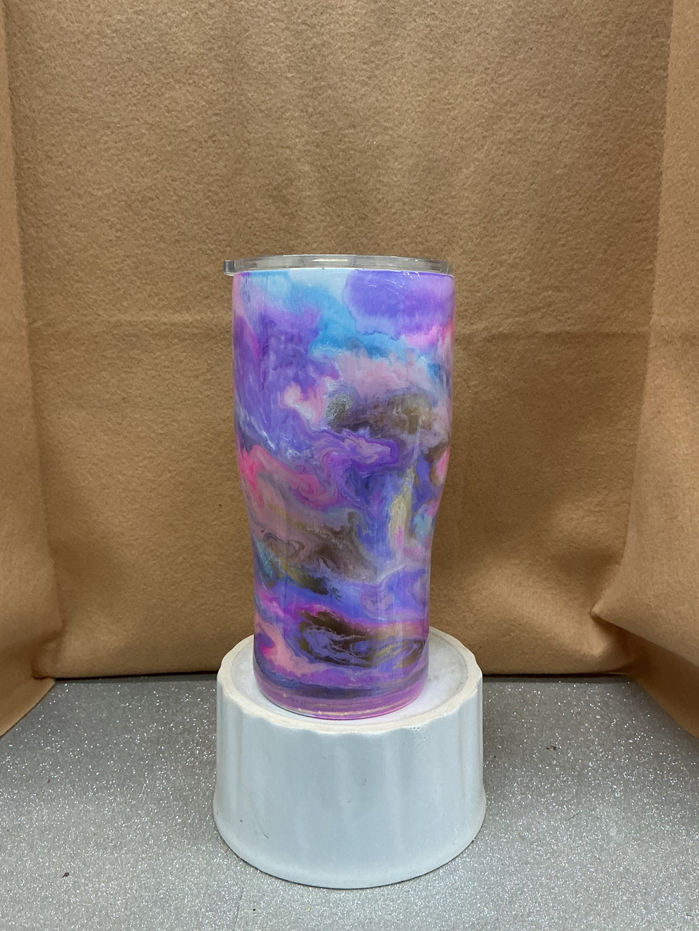 Tall cylindrical tumbler with a marbled dark pastel purple, pink, blue, and black design, placed on a white pedestal against a brown background.